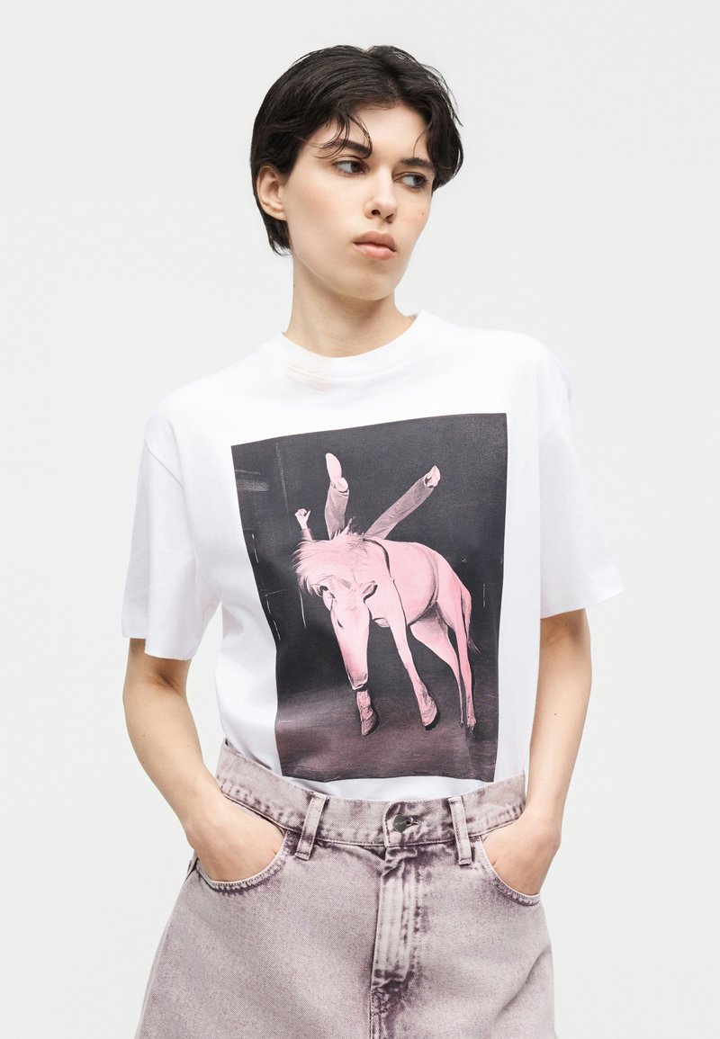 Persona con capelli corti e scuri che indossa una maglietta bianca con grafica di un cavallo rosa e ha le mani nelle tasche dei pantaloni rosa chiaro a vita alta, guarda di lato.