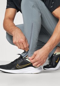 Harmaat urheiluhousut, joissa on vetoketjulliset lahkeet, korostaen sileää pintaa. Mustat ja kultaiset Nike-juoksukengät, joissa on verkkopinta ja valkoiset pohjat.