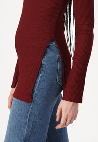 Burgundska teksturirana majica dugih rukava s bočnim prorezima, u kombinaciji s svijetloplavim trapericama. Outfit sadrži rebrasti dizajn pletenine.