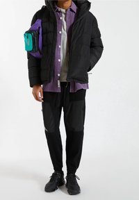 Personne portant une veste matelassée noire, une chemise violette à boutons par-dessus un t-shirt gris, un pantalon de jogging noir, des chaussures noires, et un sac à bandoulière violet avec une pochette turquoise.