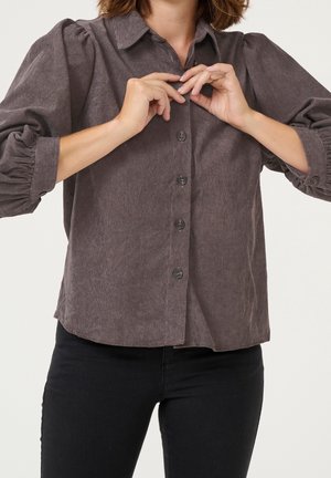 Personne boutonnant une chemise en velours côtelé gris à manches longues, portant un pantalon noir, sur un fond clair uni.