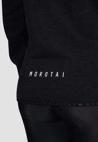 Schwarzes Sportoberteil mit einem weißen "MOROTAI"-Logo auf dem unteren Rücken, aus weichem Stoff mit glatter Textur und figurnaher Passform.