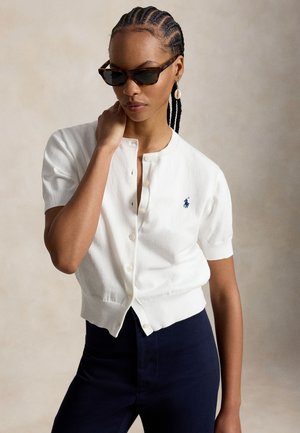 Γυναίκα με πλεγμένα μαλλιά που φορεί γυαλιά ηλίου με πλαίσιο από χελώνα, λευκή ζακέτα με κουμπιά και λογότυπο, σκουλαρίκι σε σχήμα κοχυλιού και σκούρο παντελόνι, ποζάροντας σε ουδέτερο φόντο.