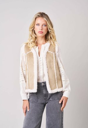 Femme blonde aux yeux bleus portant un gilet en peau retournée beige par-dessus un chemisier blanc et un jean gris taille haute, debout devant un fond uni.