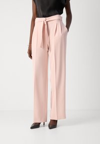 Pantaloni a gamba larga rosa chiaro realizzati in tessuto morbido, con vita alta e cintura legata, dotati di tasche plissettate. Abbinati a tacchi neri.