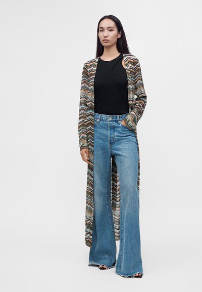 Femme portant un long cardigan multicolore, un top noir sans manches, un jean bleu taille haute à jambes larges, debout avec une main dans la poche.