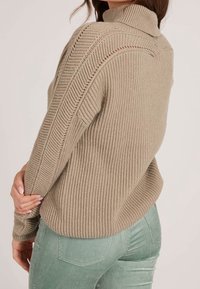 Pull beige en maille côtelée avec col montant, présentant des motifs tricotés diagonaux distincts et des manches longues, associé à un pantalon en velours côtelé menthe.