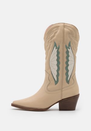 Botte de cowboy en cuir beige avec un bout pointu, un talon bloc, et un motif brodé vert et blanc en forme d'ailes sur la tige.