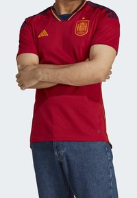 Červený sportovní dres s krátkými rukávy a výstřihem do V, s žlutými akcenty, týmovým znakem a diagonálními modrými pruhy na ramenou.