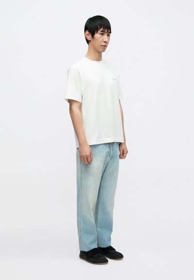 Λευκό βαμβακερό T-shirt με άνετη εφαρμογή και μικρό λογότυπο, συνδυασμένο με ξεθωριασμένα τζιν σε ανοιχτό γαλάζιο και μαύρα παπούτσια slip-on. Στάση μπροστά σε ουδέτερο φόντο.