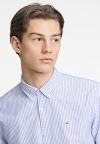 Camicia azzurra chiara a righe bianche con colletto. Presenta un piccolo logo sul petto e bottoni bianchi sul davanti.