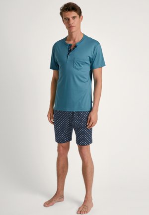 Teal short-sleeve shirt with a pocket, featuring a patterned blue neckline; paired with navy shorts adorned with white square patterns.