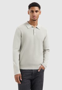 Helles graues Strick-Polohemd mit einer gerippten Textur, zwei Knöpfen und einem weichen Kragen. Modell trägt es mit dunklen Jeans.