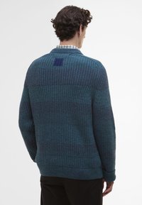 Μπλε πλεκτή ζακέτα με υφή λωρίδων, ριγέ μανσέτες και τετράγωνη λεπτομέρεια κολλάρου στο επάνω μέρος της πλάτης. Απλός σχεδιασμός και ελεύθερη εφαρμογή.