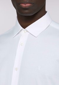 Biała koszula z długim rękawem i kołnierzykiem typu spread, wykonana z gładkiego materiału z delikatną teksturą. Zawiera pojedynczy haftowany logo na klatce piersiowej.