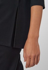 Zwarte gestructureerde stoffen top met een zijsplit, voorzien van korte mouwen en een close-up van een hand die ernaast rust.