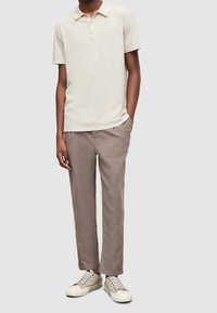 Polo beige clair avec col et trois boutons, associé à un pantalon taupe et des baskets claires. Le tissu semble lisse avec une coupe décontractée.