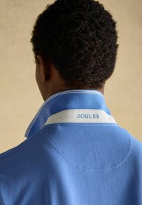 Persoană cu păr scurt și creț poartă un tricou polo albastru cu guler ridicat, având brodat pe o bandă albă textul „JOULES”.