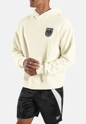 Man draagt een crèmekleurige Reebok-hoodie met voorzak en zwarte Reebok-short met witte streep, staande met gevouwen handen.