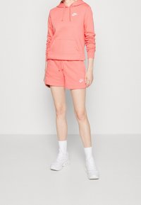 Růžová mikina s kapucí a klokaní kapsou, doplněná o odpovídající kraťasy. Obě obsahují bílé logo Nike. Model má na sobě bílé ponožky a boty.