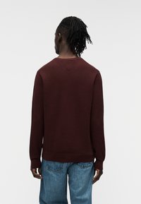 Μπορντό πλεκτό πουλόβερ με μακριά μανίκια, στρογγυλή λαιμόκοψη και υφασμένο μοτίβο· συνδυασμένο με ανοιχτόχρωμο μπλε τζιν παντελόνι.