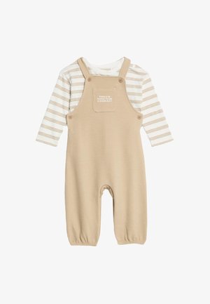 Salopette beige a coste con polsini elasticizzati e bottoni sulle bretelle, indossata sopra una maglietta a maniche lunghe bianca e beige a righe con testo.