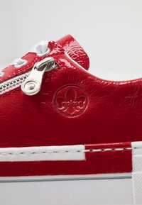 Baskets en cuir verni rouge avec lacets blancs et une fermeture éclair argentée. Présente un logo en relief et une surface texturée, avec une semelle blanche contrastante.