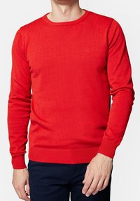 Pull en tricot rouge avec un col rond et des poignets côtelés. Présente une texture lisse et un petit logo sur la poitrine.