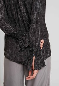 Blusa nera a trama con lucentezza metallica, caratterizzata da polsini arricciati e dettagli con bottone. Abbinata a pantaloni grigi chiari, dal taglio morbido.