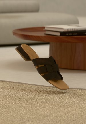 Sandale mule noire tissée avec un petit talon carré et une semelle intérieure lisse couleur tan, présentée au-dessus d'un tapis beige texturé et d'une table basse en bois.