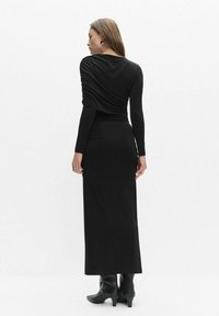 Czarna sukienka z długim rękawem o drapowanym fasonie na plecach; wykonana z gładkiego materiału. Ma dopasowany krój i jest zestawiona z czarnymi botkami na obcasie.