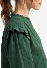 Green top crafted from textured fabric featuring vertical stripes, ruffled sleeve edges, and a high neckline.
