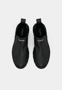 Μαύρα παπούτσια slip-on από λεία ύλη, με υφή στη σόλα, στρογγυλή μύτη και επώνυμη λεπτομέρεια στην άνω επιφάνεια.
