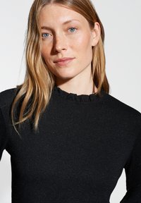 Schwarzes Longsleeve-Oberteil aus strukturiertem Stoff, mit einem Rüschenausschnitt und dezentem Glitzer. Enganliegendes Design ohne sichtbare Verschlüsse.