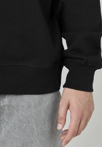 Sweatshirt noir avec des poignets et un ourlet côtelés, présenté sur un tissu gris clair. La texture est lisse et légèrement extensible.