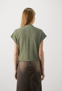 Olivgrünes Kurzarmoberteil mit lockerer Passform, strukturierter Textur und abgerundetem Saum. Kombiniert mit einem dunkelbraunen Rock mit einer hinteren Naht.