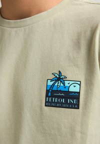 Lichtgroene katoenen t-shirt met een rechthoekig blauw en zwart logo met een palmboom, oceaangolven en de tekst "PETROL IND." in zwart.