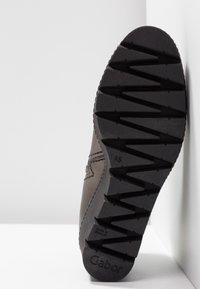 Semelle d'une chaussure Gabor marron avec un motif de bande de roulement en zigzag profond, inclinée verticalement contre un fond blanc uni.