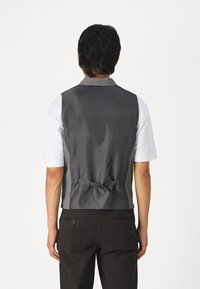 Gilet grigio con retro testurizzato, cravatta regolabile e pannello frontale liscio. Abbinato a una camicia bianca e pantaloni scuri. Design semplice.