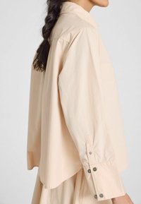 Camicia oversize beige chiaro con colletto a punta e maniche lunghe. Presenta cuciture a vista e bottoni ai polsini.