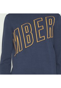 Camiseta de manga larga en color marino con un texto bordado en amarillo que dice "MBER" en la parte frontal. Tela suave con un ajuste regular.