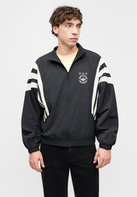 Chaqueta negra con cremallera y detalles en rayas blancas, logo en el pecho, puños y borde elásticos, y tejido ligero. Se lleva sobre una camisa clara.