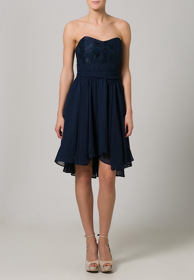 Femme portant une robe bleu marine sans bretelles jusqu'aux genoux avec un corsage en dentelle et une jupe fluide, associée à des sandales beiges à talons hauts.