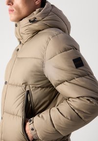 Veste rembourrée beige avec un col haut, une poche zippée et un patch logo noir sur la manche. Présente une texture lisse et brillante.