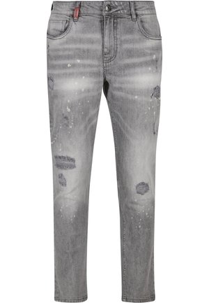 Mann steht in grauen zerrissenen Jeans und weißen Sneakers mit schwarzen Sohlen vor einem schlichten weißen Hintergrund, Seitenansicht.