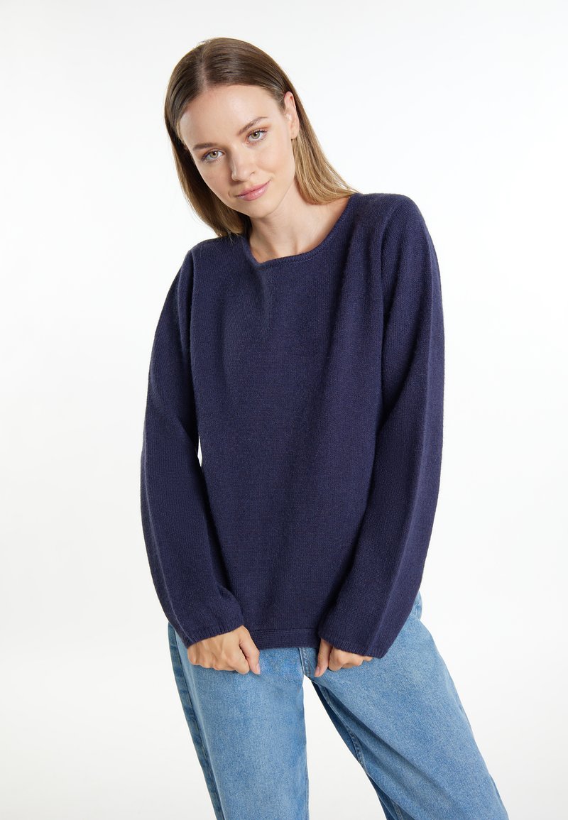 Marineblaues Strickoberteil mit weit geschnittenem Ausschnitt und langen Ärmeln, hergestellt aus einem weichen, strukturierten Material. Kombiniert mit hellblauen Jeans.
