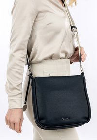 Borsa a tracolla in pelle nera con finitura texturizzata, dotata di una tracolla removibile e accessori in metallo color argento, tenuta da una persona che indossa una blusa beige.