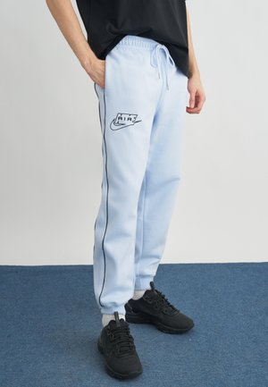 Asmuo dėvi šviesiai mėlynas Nike Air sportines kelnes su juoda šonine juostele ir juodus sportinius batelius, stovintis ant mėlyno kilimo.