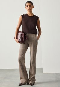 Top marrone senza maniche abbinato a pantaloni larghi a quadri in tonalità terrose. Accessori includono una borsa marrone scuro e scarpe marroni a punta.