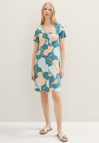 Frau in einem kurzärmeligen, V-Ausschnitt-Kleid mit großen blauen und pfirsichfarbenen Blumenmustern, barfuß auf einem hellen Hintergrund stehend.